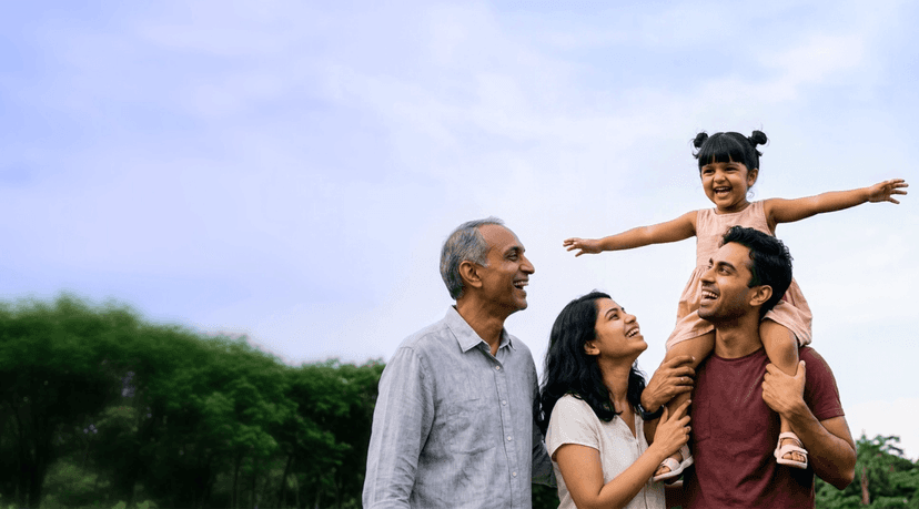 Three generations of an Indian family laughing together outdoors, with a young girl on her father’s shoulders